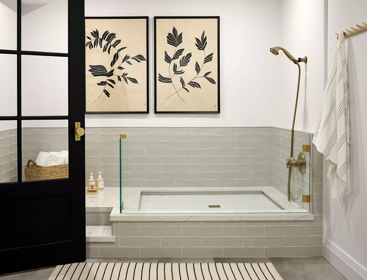 White bathroom with a black door. There is a large tub set up to bathe dogs it has grey tiles. There are two black and white prints above the bath.